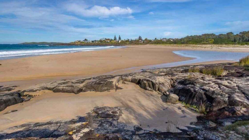 Sandy beach with rocky foreground and ocean waves under a blue sky.