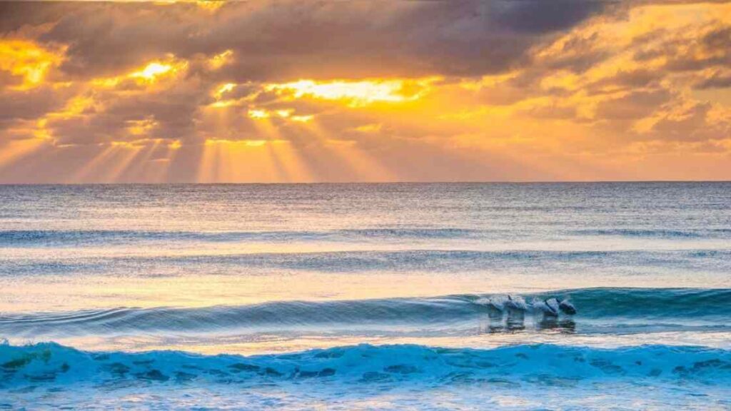 Sunrise over the ocean with golden rays shining through clouds and gentle waves.